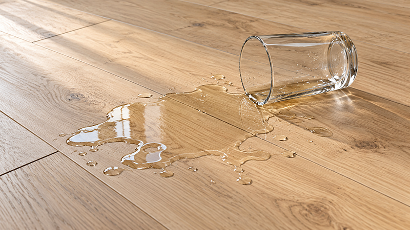 Umgekipptes Wasserglas mit Wasserpfütze auf wasserresistentem Laminat in Eichenoptik